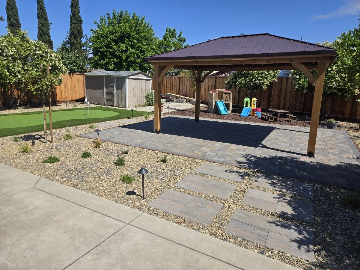 Backyard with a covered patio, small artificial grass putting green, children's play area with slides and sandbox, wooden fence, garden beds, small trees, and pathway with stone tiles under bright sunny sky.
