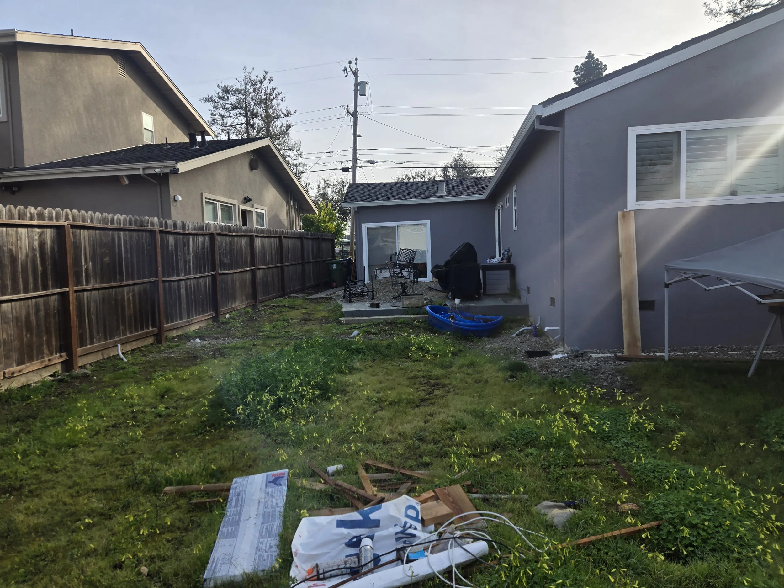 Backyard scene with two houses, a wooden fence, outdoor furniture, a barbecue grill, grass, and scattered debris including a boxed item, plastic bags, and miscellaneous materials.