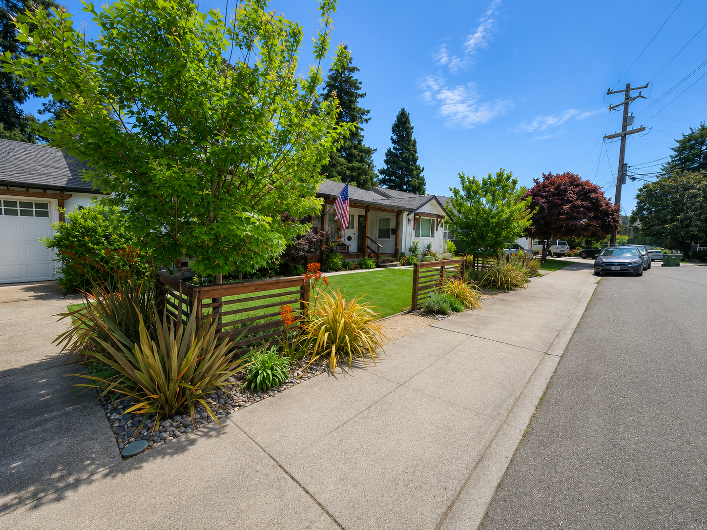 A suburban street with a single-story house, lush front yard with various trees and plants, a small fenced garden, parked cars, utility poles, and a clear blue sky.