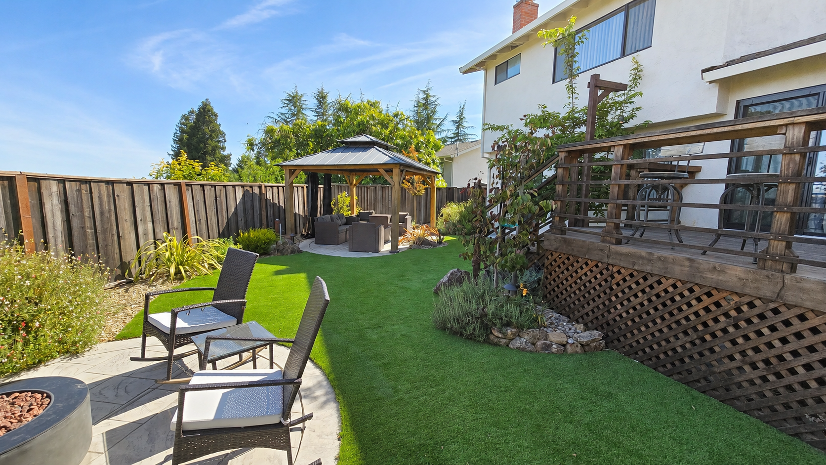 Backyard with a wooden fence, a grassy area, outdoor seating beneath a gazebo, and a deck with chairs and tables, in front of a white house.