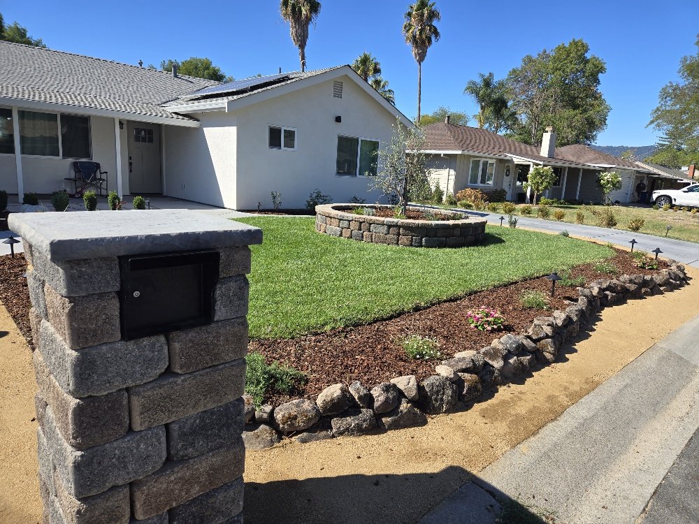 Front yard with a manicured lawn, circular raised flower bed, and landscaped border with rocks, in front of a white house with a grey roof, under a clear blue sky with palm trees.