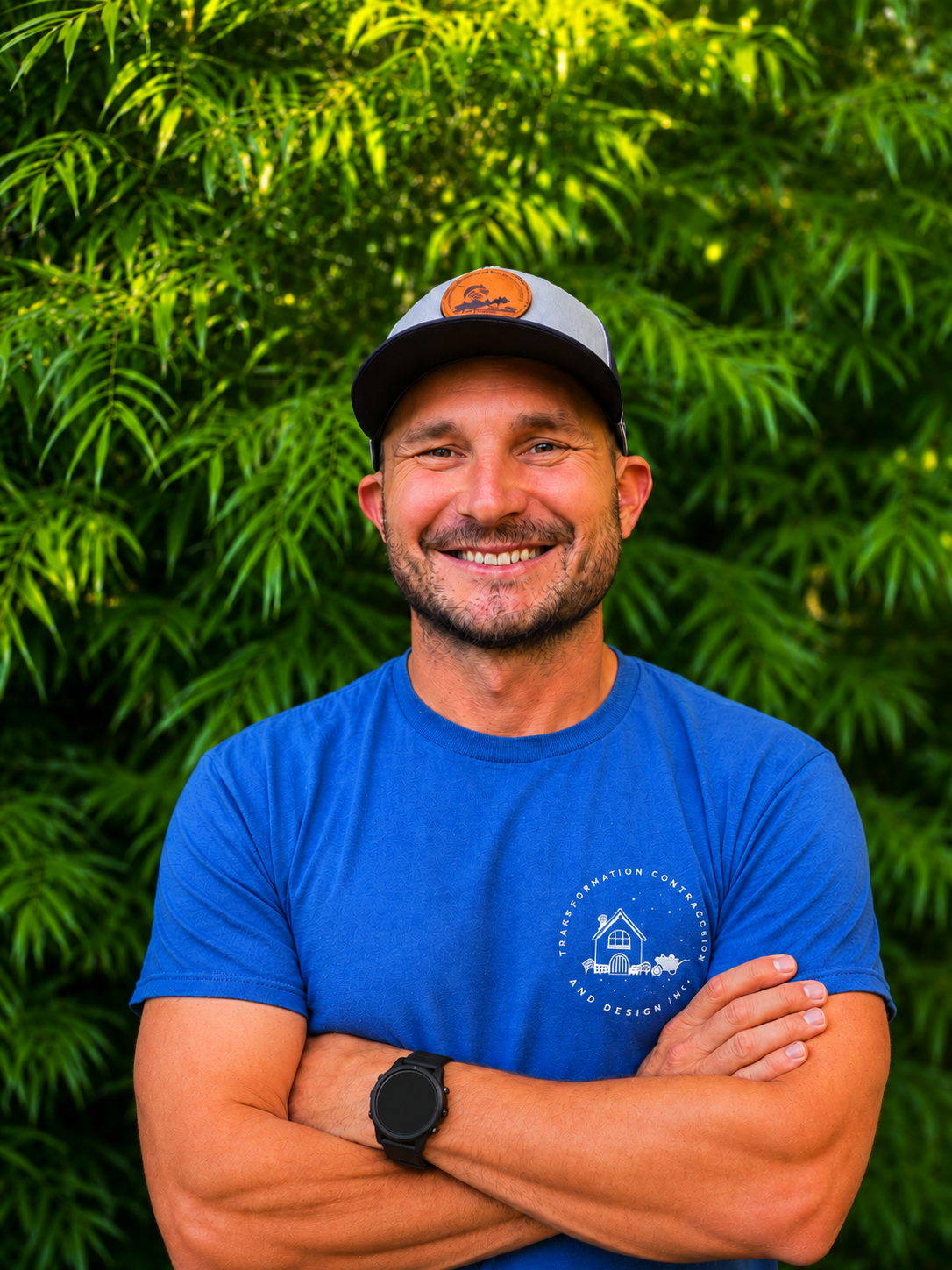 A man wearing a hat and a blue shirt stands, smiling at the camera