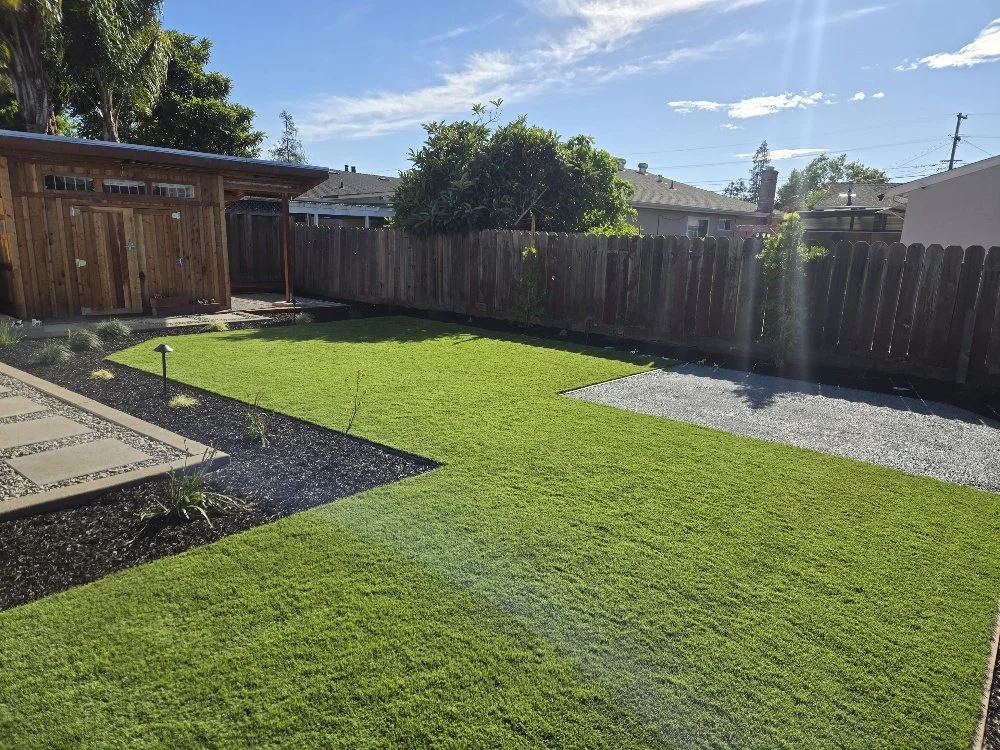 A backyard with a well-maintained lawn, a small garden bed with plants, a wooden shed, a gravel area, and a wooden fence surrounding the yard under a sunny sky.