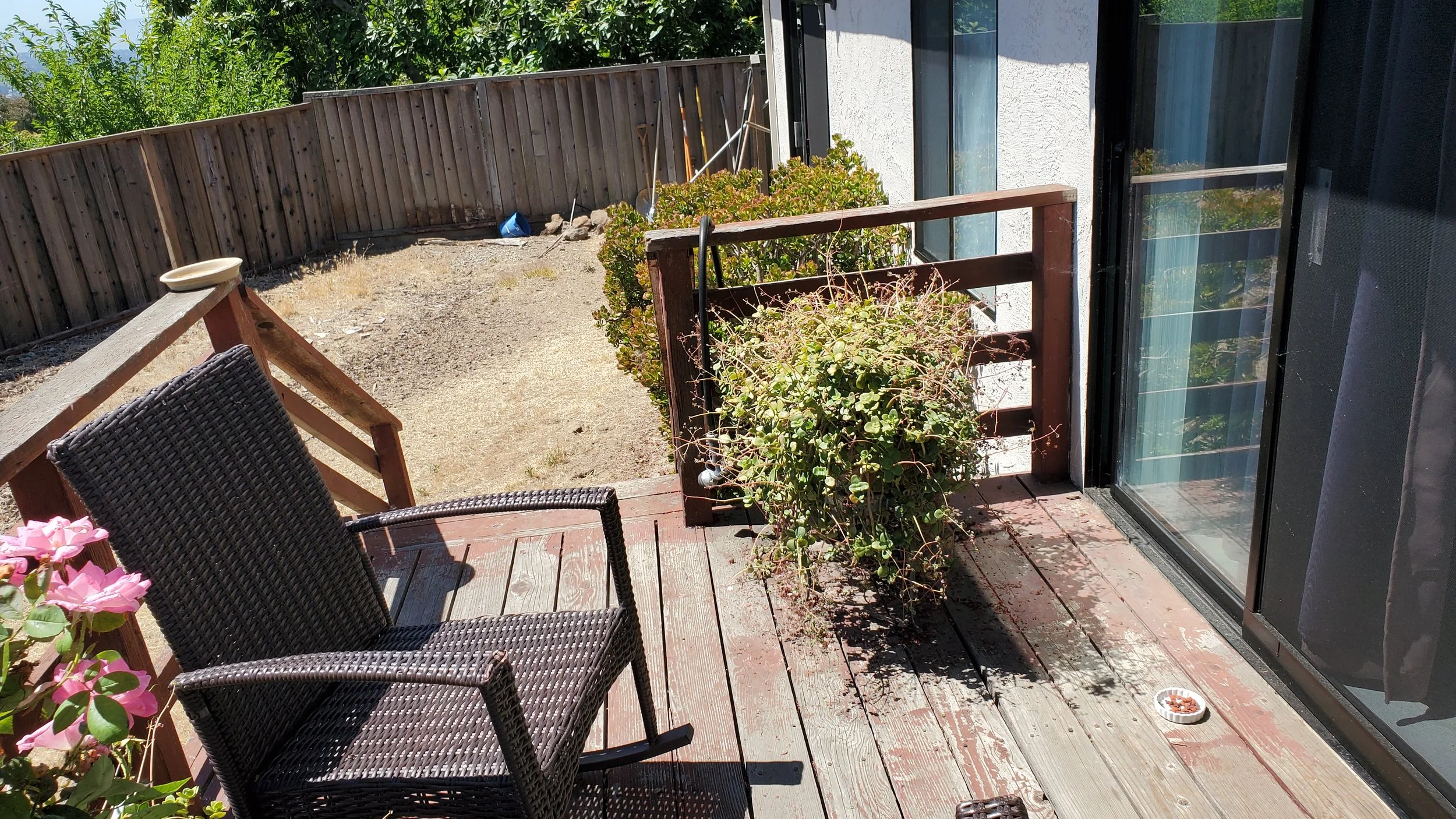 A backyard patio with wooden decking, black wicker chairs, pink flowers, potted plants, and a fenced yard with shrubs and trees.