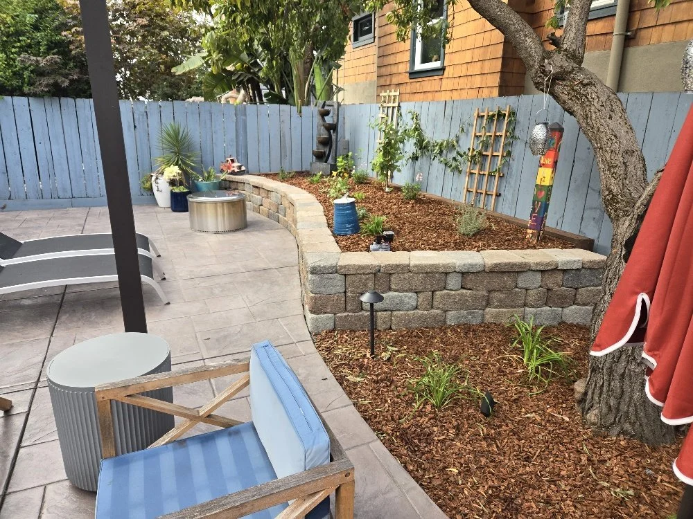 Backyard patio with a blue fence, potted plants, outdoor furniture, and a garden bed with plants and decorations.