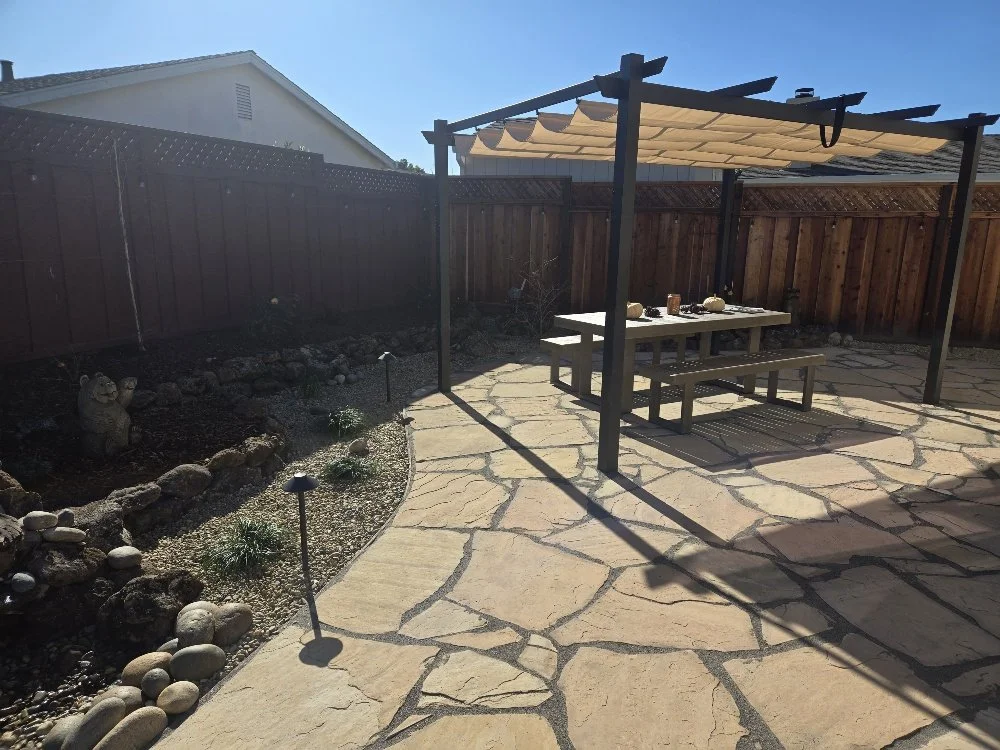 A backyard patio with a wooden picnic table and benches under a pergola with a fabric shade, surrounded by a stone pathway, a garden bed with rocks and plants, and a brown wooden fence.