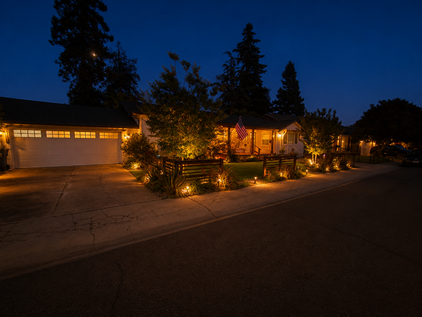 Night view of suburban houses with warm outdoor lighting, trees, and an American flag on the front porch.