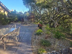 An outdoor patio area with metal chairs and a table, surrounded by trees and greenery, with a stone pathway and a distant house in the background.