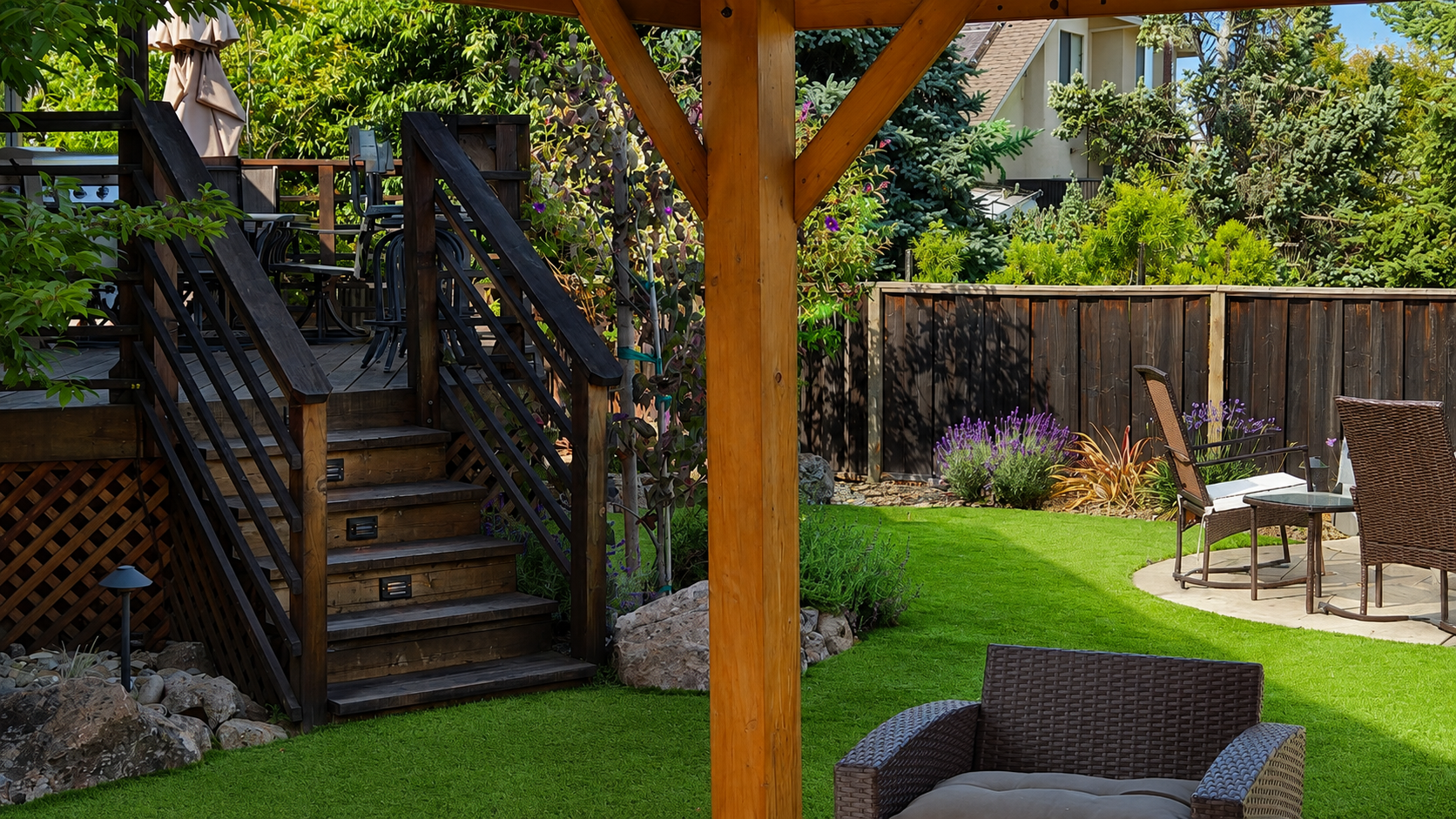 Backyard garden with patio, patio furniture, elevated wooden deck, lush green grass, trees, shrubs, and flowering plants.
