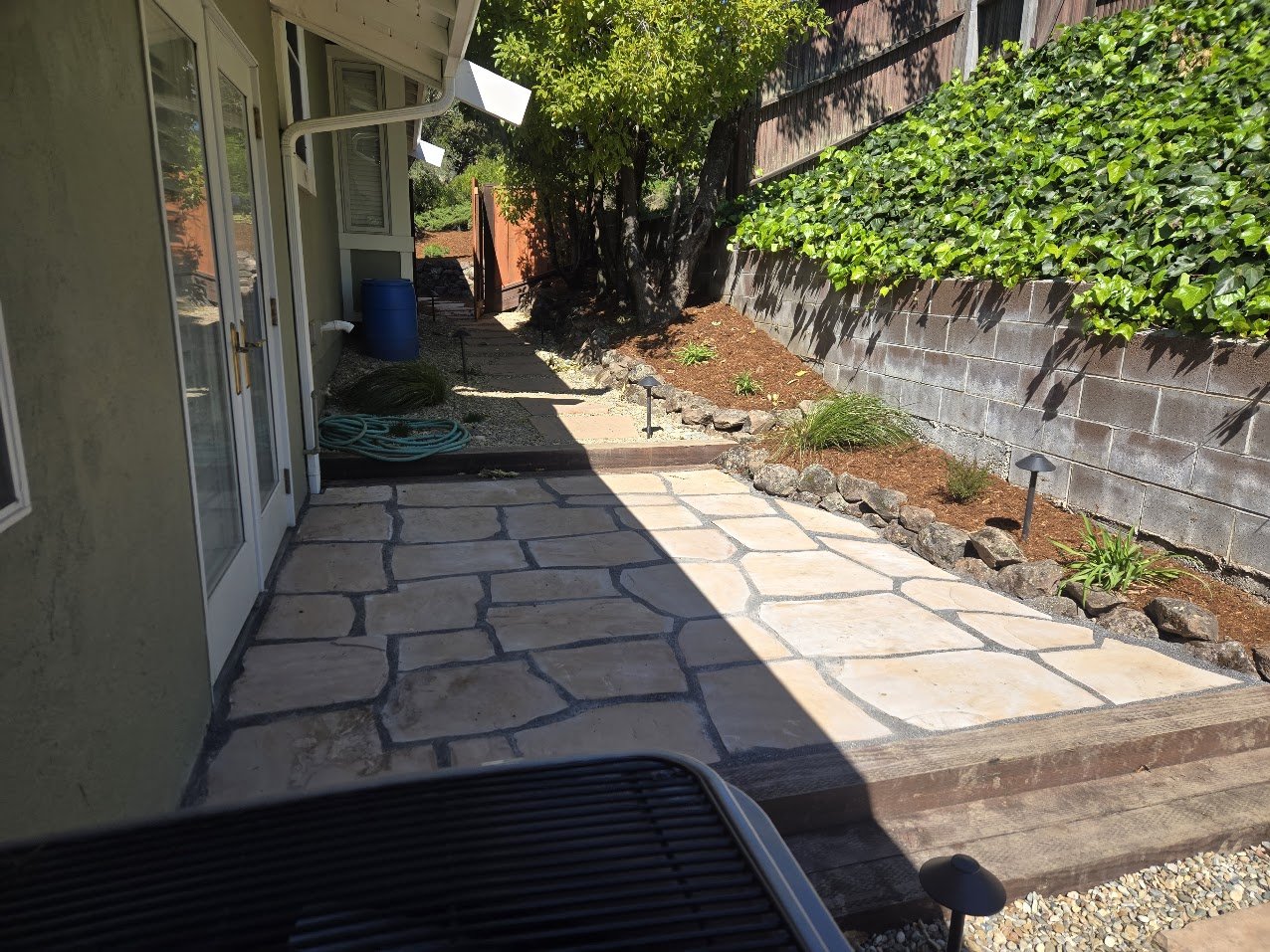 Backyard patio with flagstone flooring, garden beds with small plants, and a large tree providing shade. There are outdoor lights along the garden beds, and a concrete block wall covered with green ivy on the right side. A blue barrel and garden hose