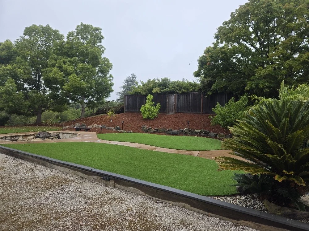 A backyard with a patch of artificial grass, a fire pit, trees, shrubs, and a wooden fence.