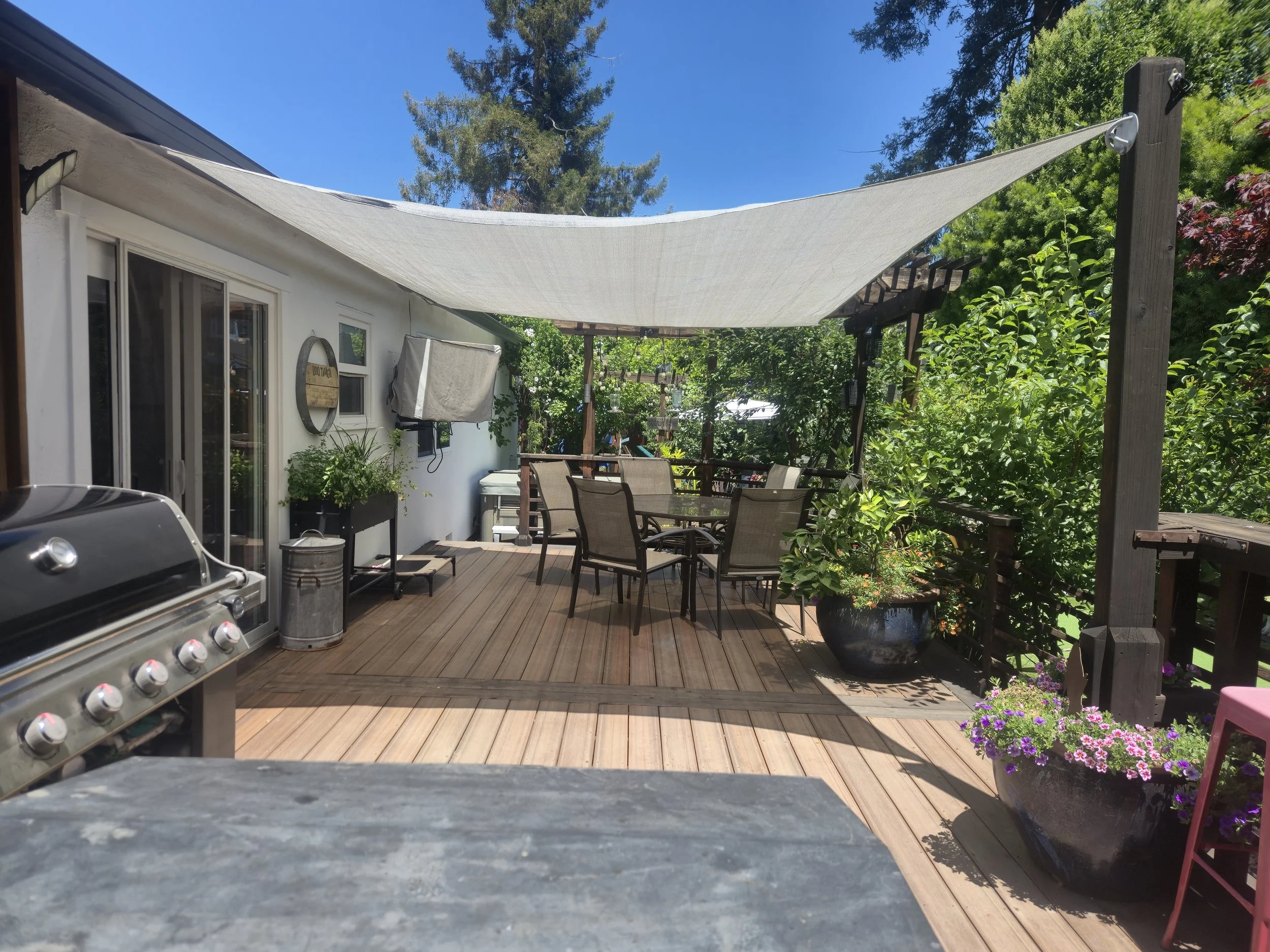 A backyard patio with a shaded area created by a large white fabric canopy, wooden deck, gardening chairs, potted plants including purple flowers, a barbecue grill, and surrounded by green trees under a clear blue sky.