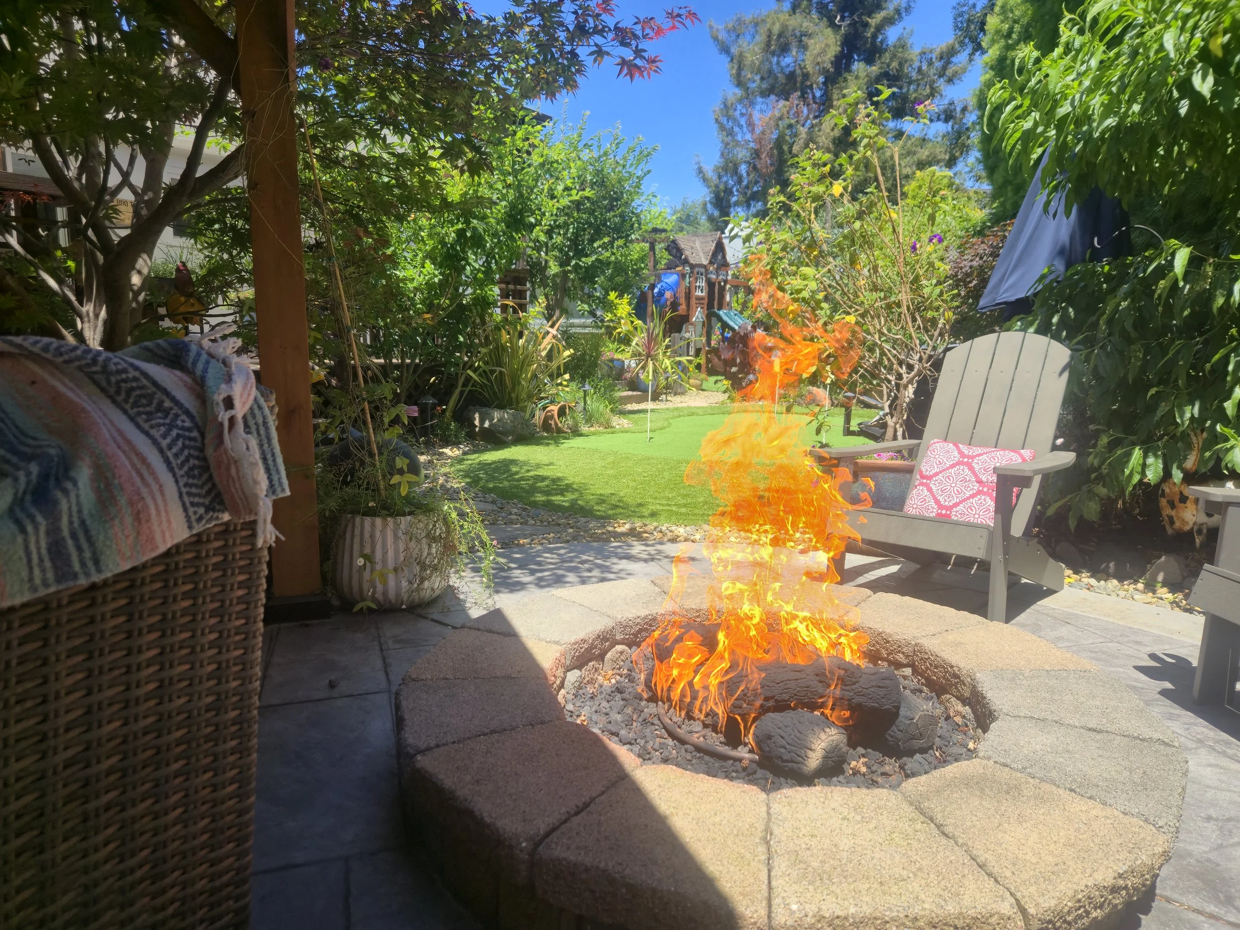 A backyard with trees, bushes, outdoor furniture, and a fire pit with flames. There are chairs, a towel draped over a basket, and clothes hanging on a line under a partly cloudy sky.