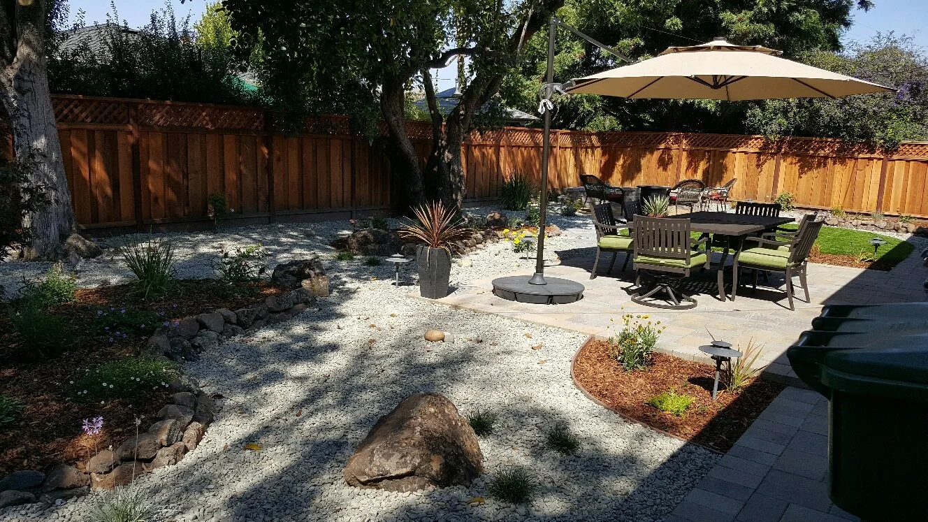 Backyard patio with wooden fence, trees, outdoor table with chairs, large umbrella, plants, and rocks on a sunny day.
