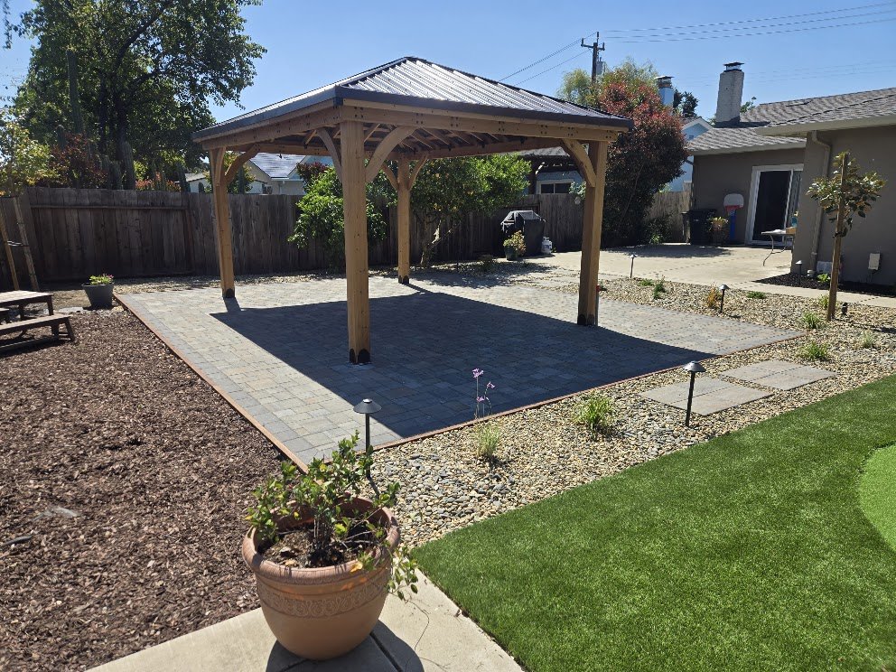 Backyard with a new wooden gazebo, a paved patio area, and a small grassy lawn surrounded by a wooden fence and plants.