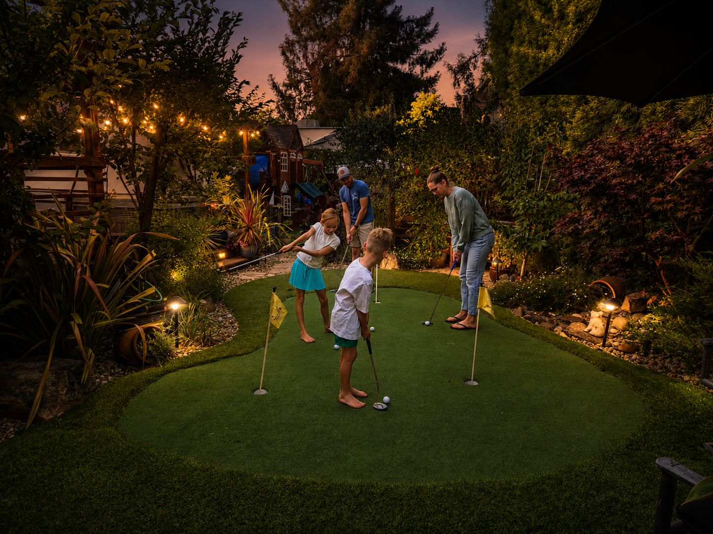Family playing mini golf on a backyard putting green during sunset, surrounded by lush plants and garden lights.