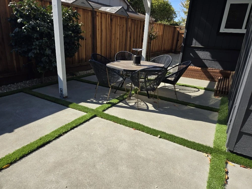 Outdoor patio with concrete and artificial grass tiles, black metal dining table and chairs, fenced yard, and a dark-colored house wall.