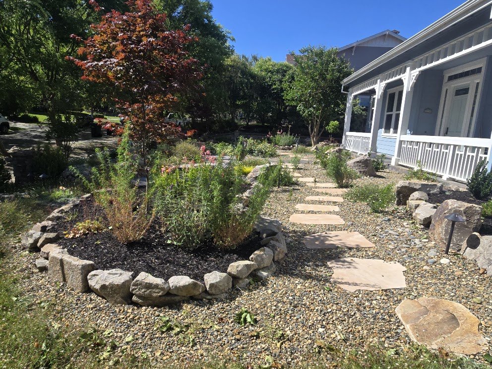 A garden pathway made of flat stones surrounded by small gravel, leading to a porch of a white house with blue accents. There are organized garden beds with various green plants, and a tree with reddish leaves to the left. The scene is sunny with a c