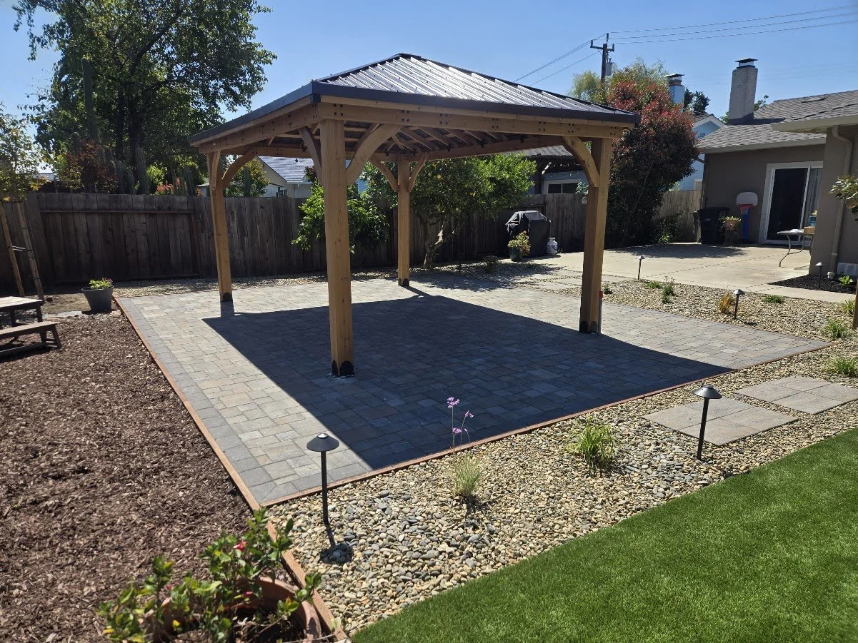 Backyard with a wooden gazebo on a paved patio, surrounded by a landscaped area with small plants, mulch, stones, and a well-maintained lawn, with house, fence, and trees in the background.