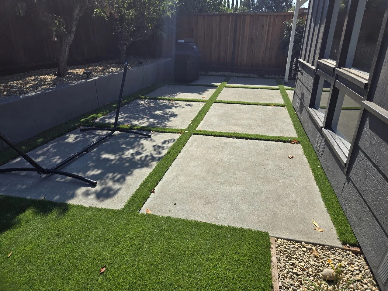 A backyard patio with concrete slabs separated by strips of artificial grass, a black grill, a wooden fence, and a neighboring house's window.
