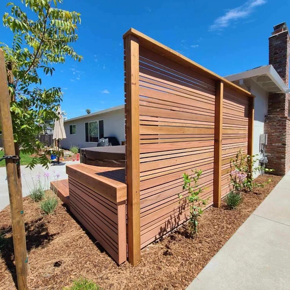 Wooden privacy fence surrounding a hot tub outside a single-story house with a brick chimney and a concrete sidewalk.