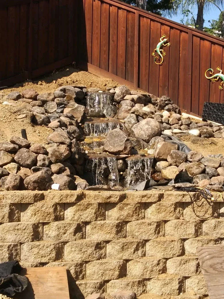 A small, three-tiered waterfall feature with rocks, situated in a backyard with a wooden fence decorated with lizard ornaments and a retaining wall of stone blocks.