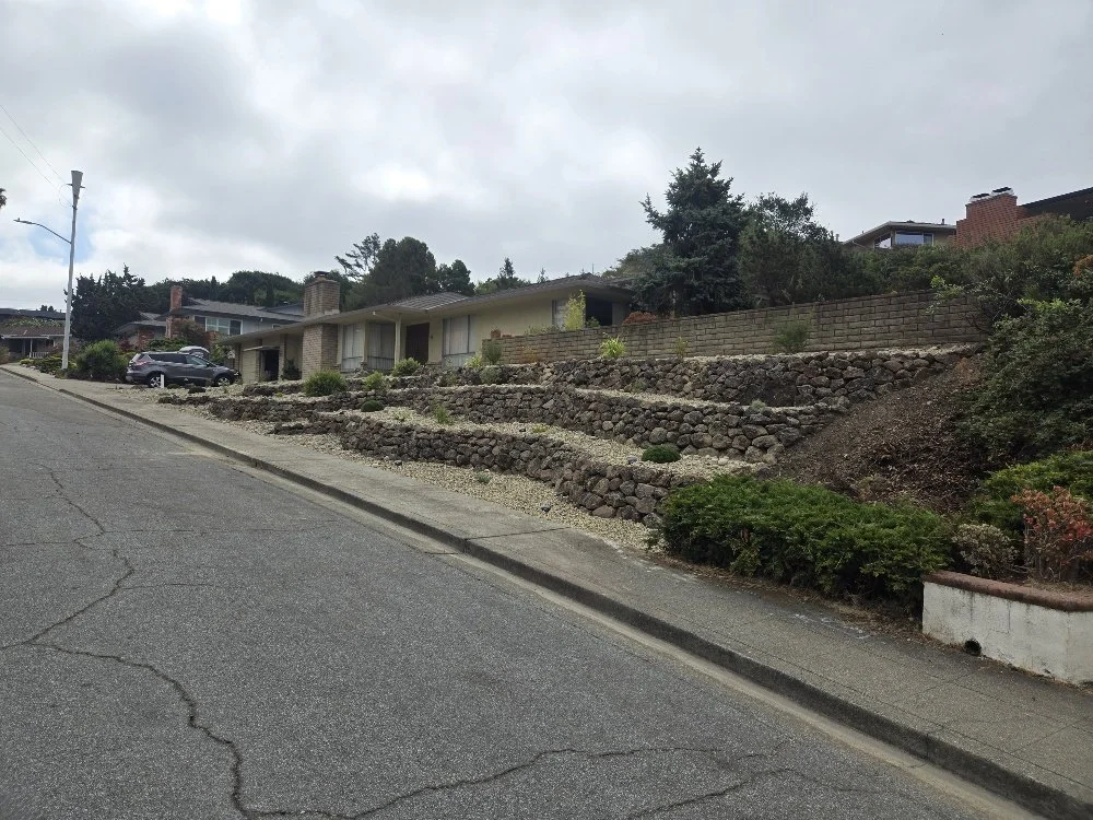 Residential neighborhood with a house on a hillside, featuring tiered rock retaining walls and some greenery, under a cloudy sky.