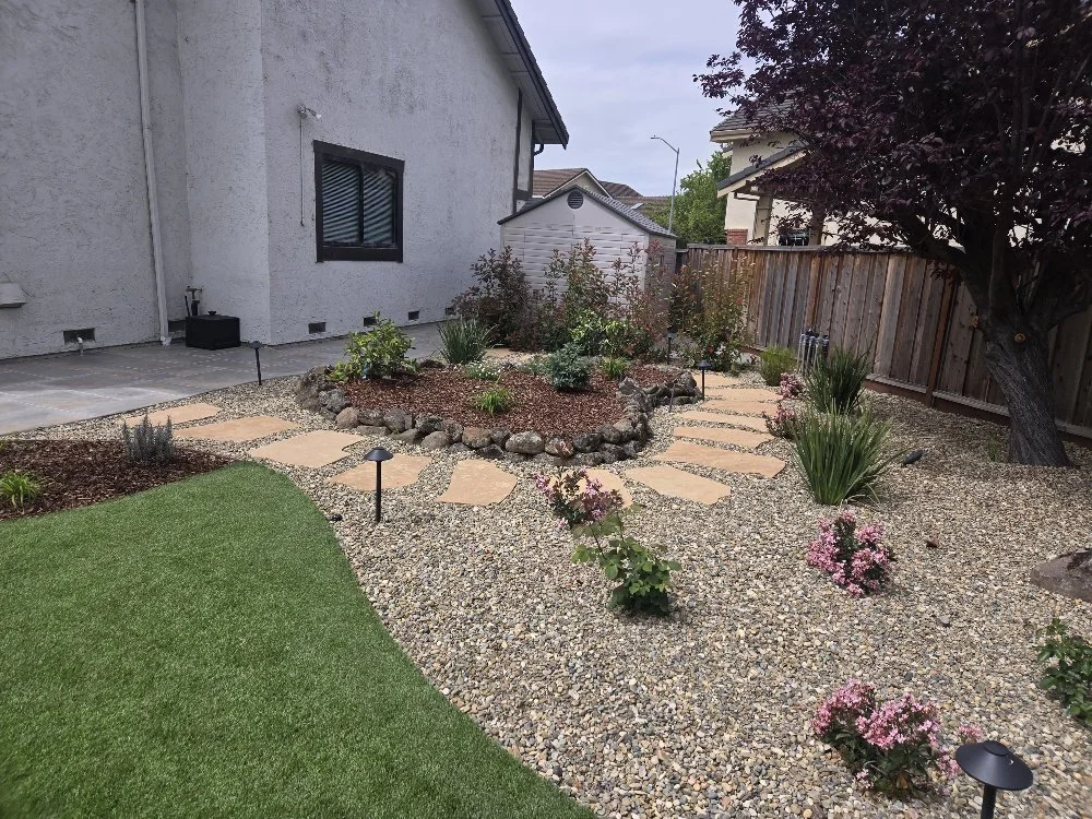 Backyard garden with pathways, flower beds, a large tree, and a wooden fence.