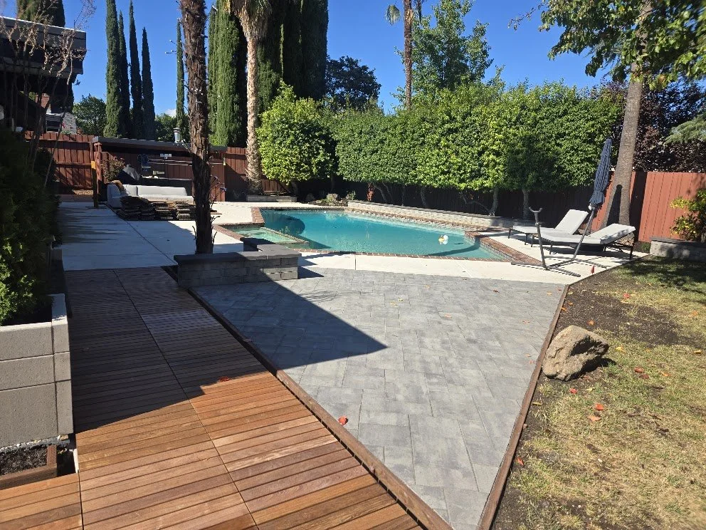 Backyard with a swimming pool surrounded by trees, greenery, and patio furniture, including lounge chairs and an umbrella, under a clear blue sky.