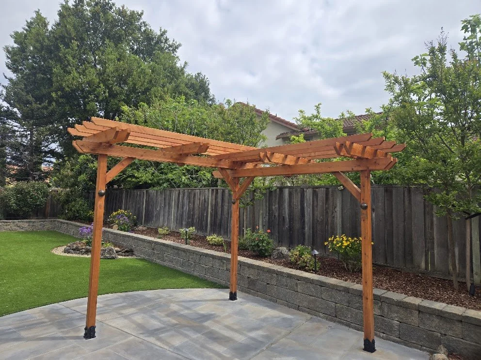 Wooden pergola with partially installed slats over a patio in a backyard garden with trees and a wooden fence.