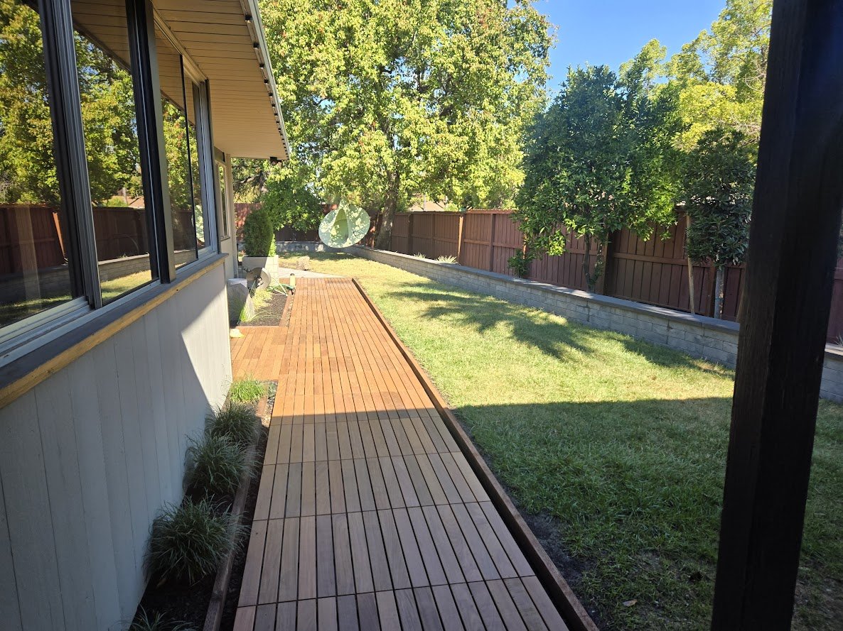 Backyard with a wooden deck along the side of a house, green grass, trees, and a wooden fence under a clear blue sky.