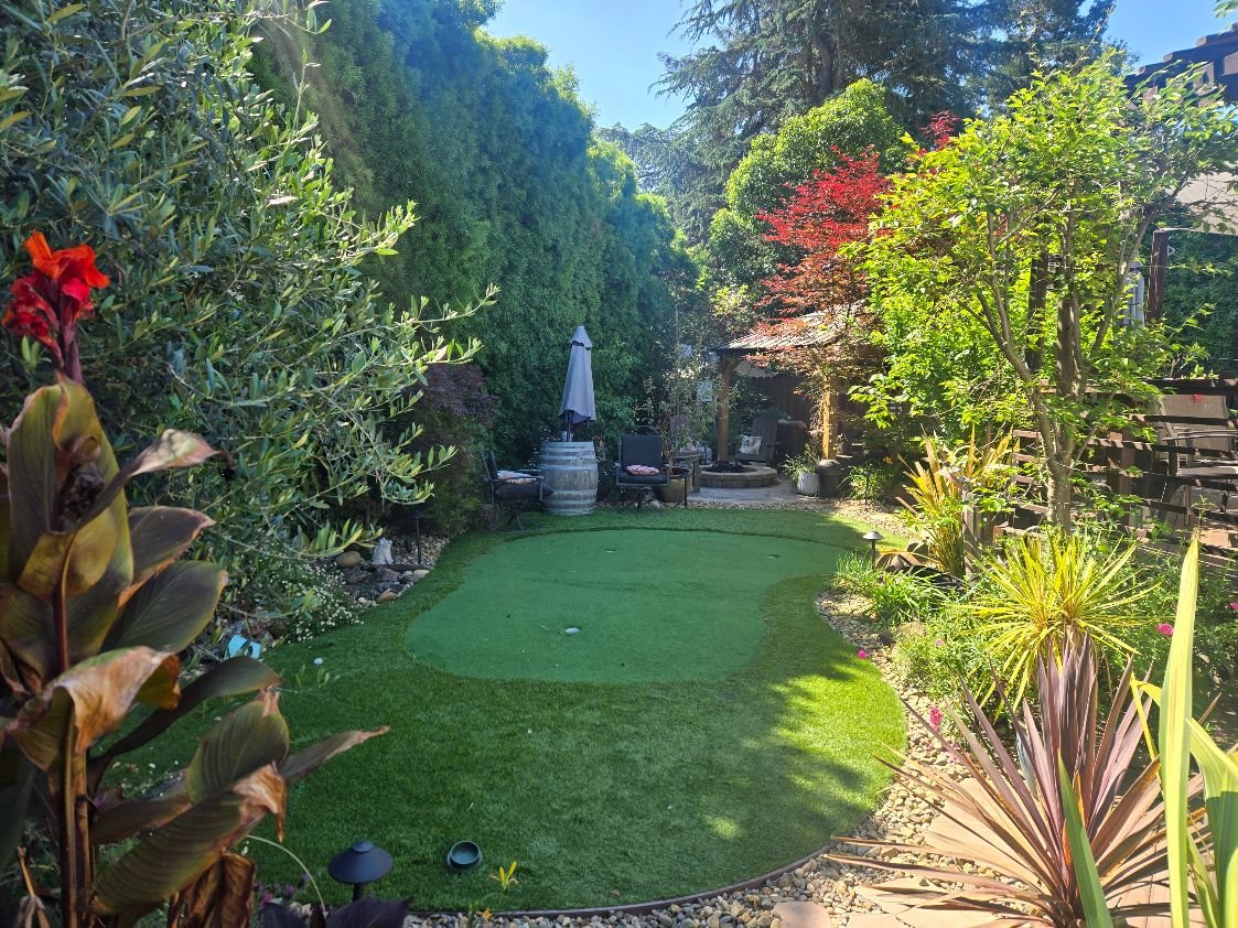 A lush backyard garden with a small putting green, surrounded by various plants and trees. There are chairs, an umbrella, and decorative barrels, creating a cozy outdoor space.