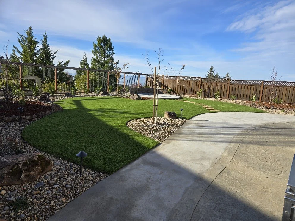 Clean yard with freshly laid green grass, small trees, and a wooden fence, with a concrete patio and pathway under partly cloudy skies.