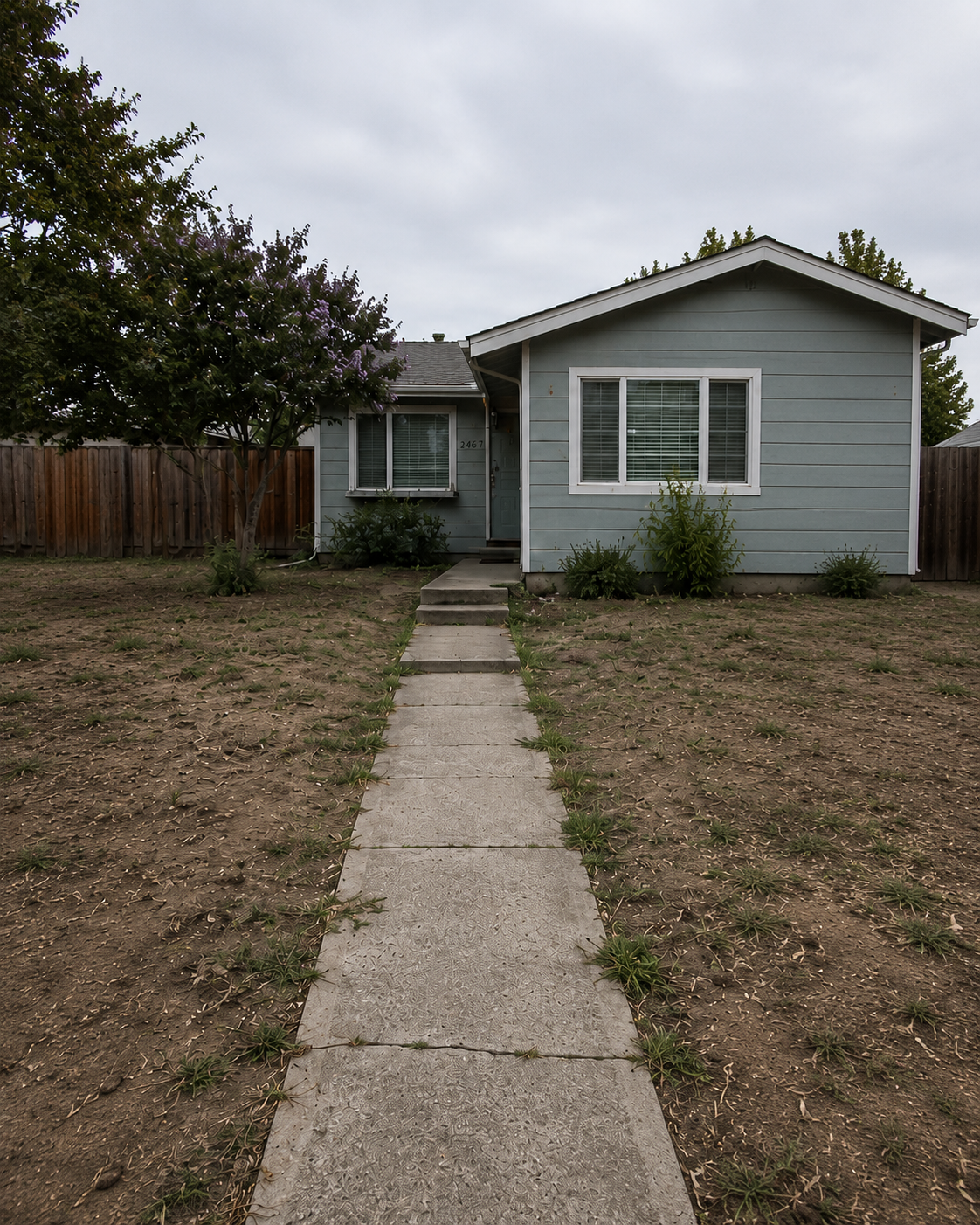 Front view of a small, light blue house with white trim and a side yard with patches of dirt and sparse grass, a concrete walkway leading to the front door, a tree with purple flowers on the left, and a wooden fence in the background under cloudy sky.