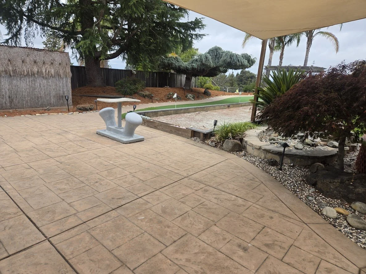 A backyard patio area with a small seating area and decorative plants. There is a putting green in the background, along with trees and a privacy fence.