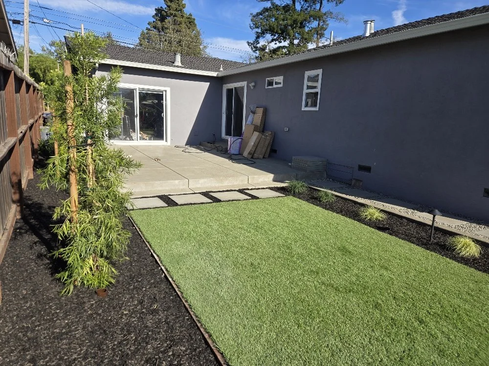 Backyard with a green lawn, concrete patio, sliding glass door, and house with gray exterior walls under a blue sky.