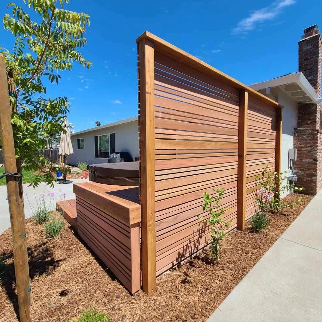 A wooden privacy fence surrounds a backyard area, with greenery and plants along the base. In the background, there is a house with a window, an outdoor hot tub, and an umbrella. The sky is clear and blue.