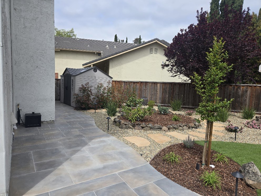 Backyard with a tiled patio, young trees, garden beds, and a wooden fence.