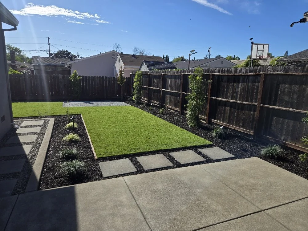 A backyard with a lawn, stone pathway, and garden beds bordered by dark mulch, surrounded by a wooden fence, under a blue sky with some clouds.