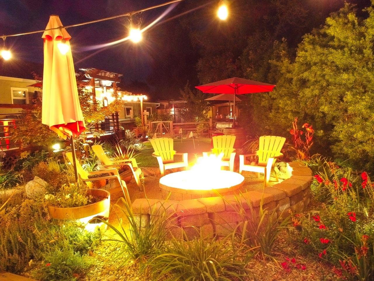 Nighttime backyard scene with a fire pit surrounded by yellow Adirondack chairs, red and yellow umbrellas, string lights, lush greenery, and colorful flowers.