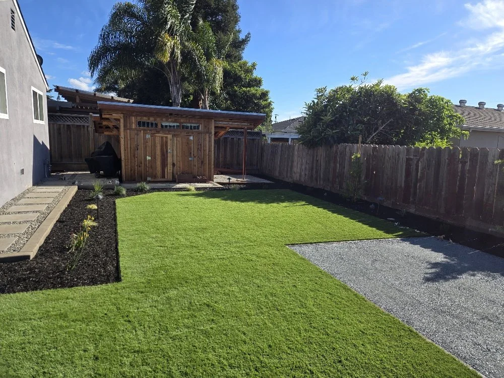 A backyard with a lawn, a small garden bed with plants, a wooden shed, a gravel area, and a wooden fence under a partly cloudy sky.
