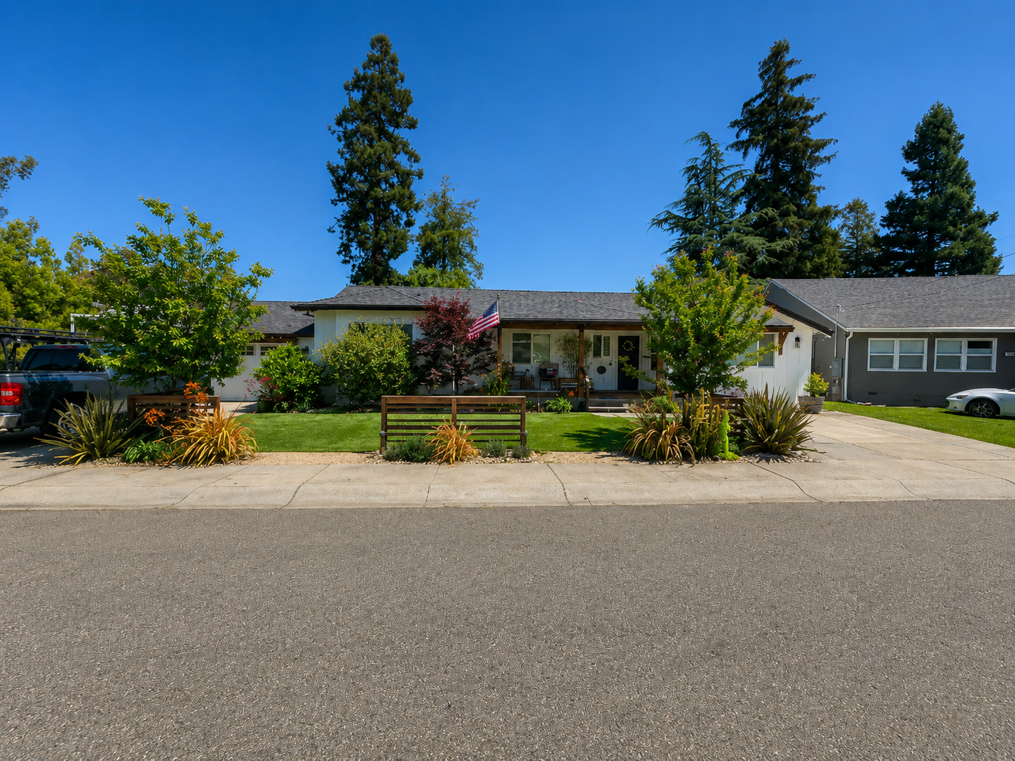 Front view of a single-story house with a well-maintained lawn, trees, and shrubs, with a driveway and parked vehicles on either side, under a clear blue sky.
