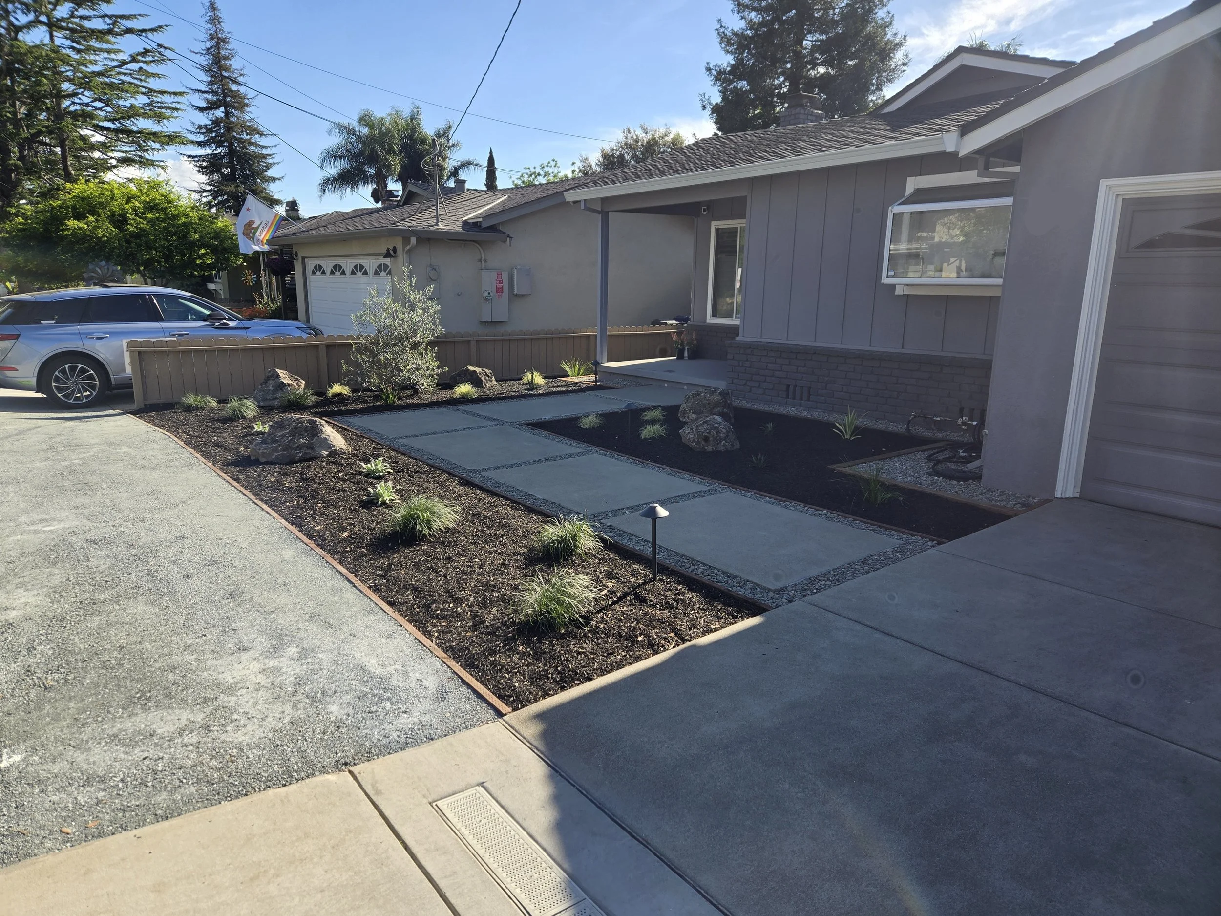 Front yard and driveway of a house with a white garage door, gray exterior walls, and a small porch with potted plants. A silver car is parked on the driveway. The landscaping includes rocks, small plants, and mulch. A California flag is visible hang