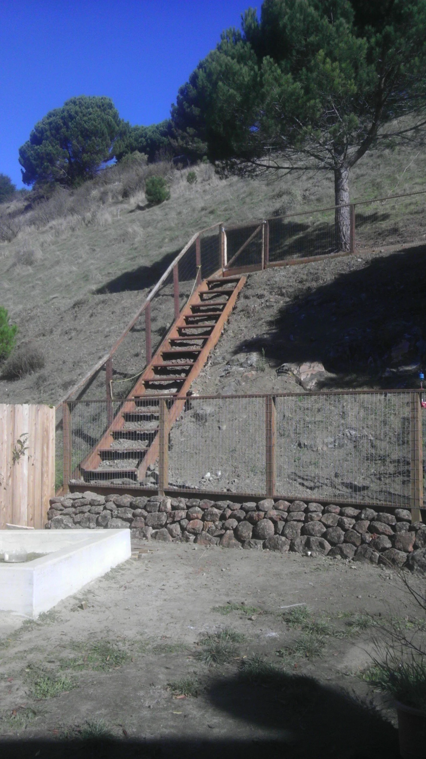 Outdoor scene with a dirt slope, a wooden staircase with wire railing, a tree, and a fence at the bottom. Clear blue sky and dry grass are visible.