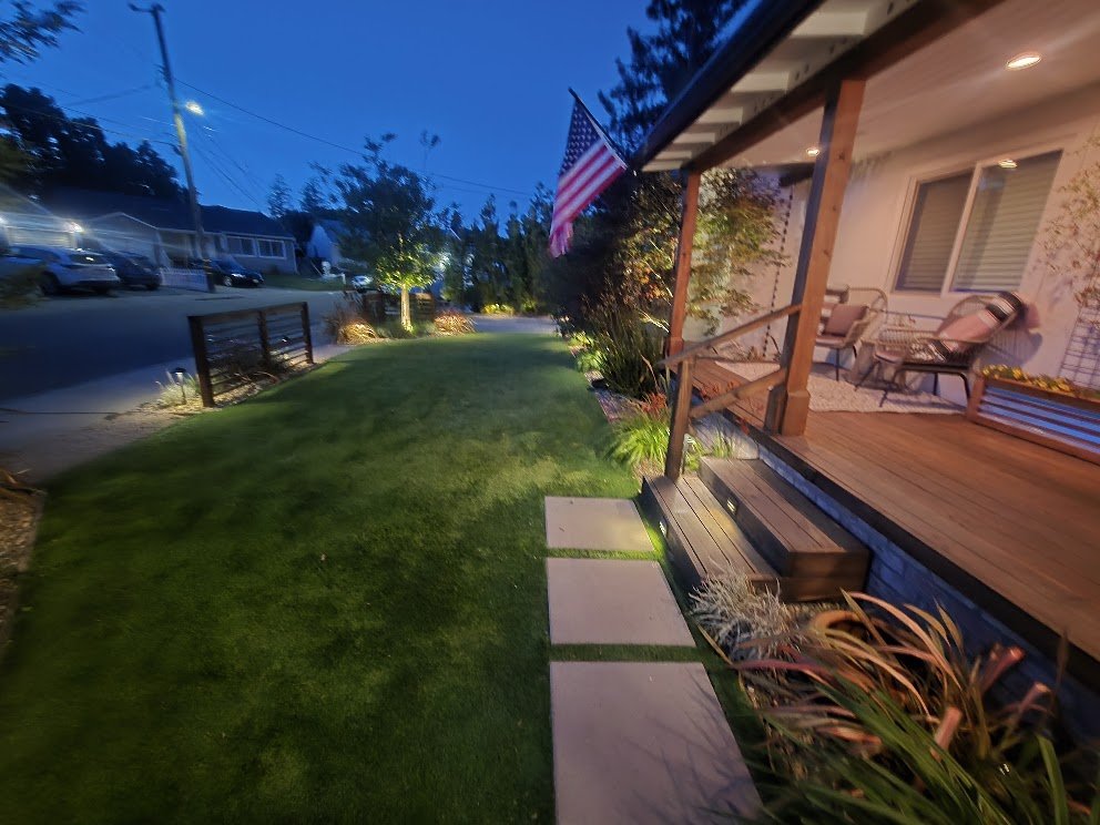 A front yard with a well-lit porch, American flag, greenery, and a stone pathway leading to the house during dusk.