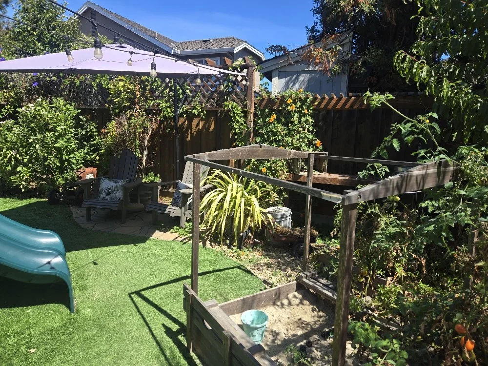 A backyard garden with a small sandpit, plastic slide, patio table with chairs under a purple umbrella, greenery, trees, and a wooden fence with flowering plants.