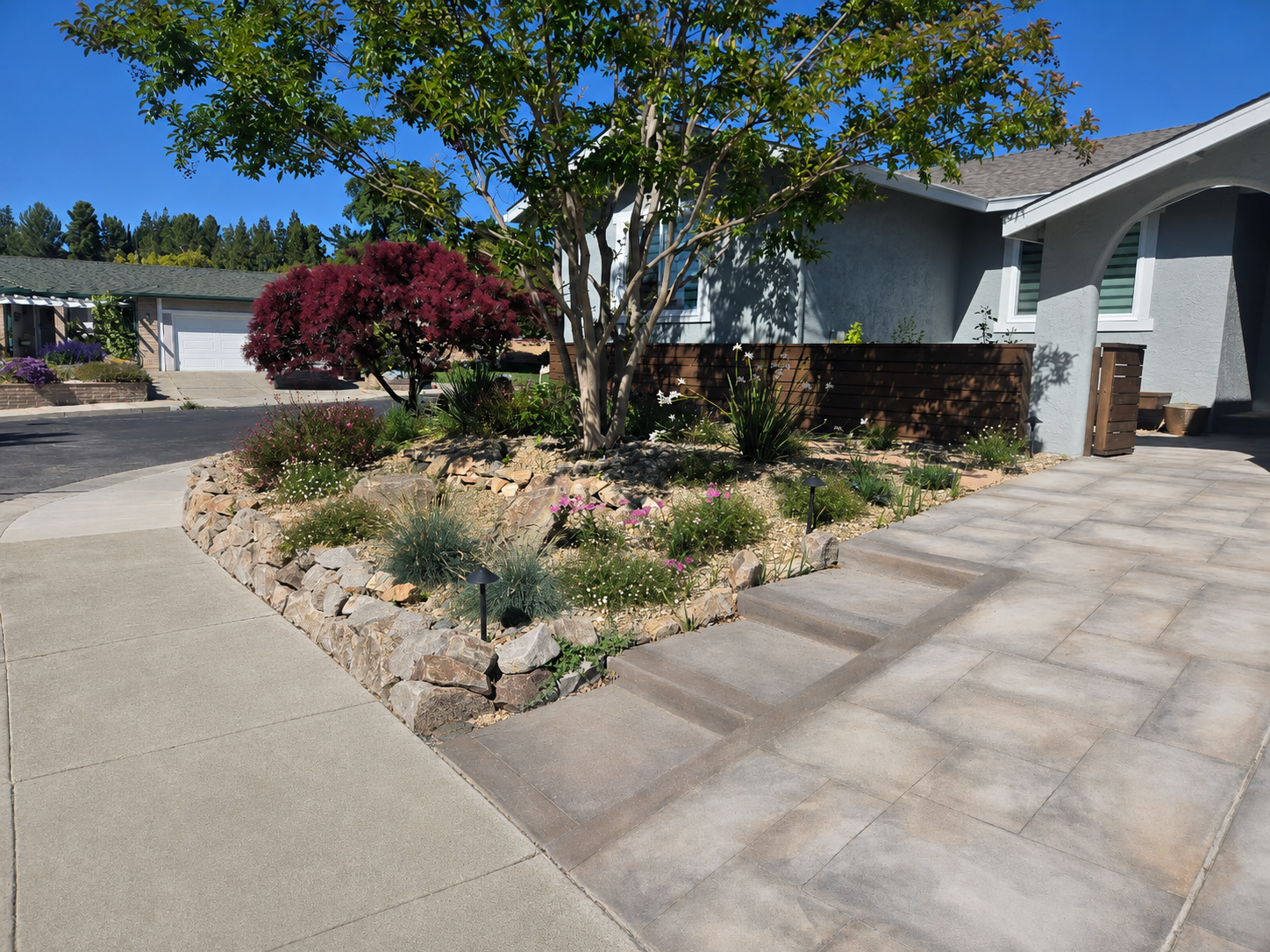 Residential front yard with landscaped garden bed featuring a small tree, flowering plants, and decorative rocks, adjacent to a paved walkway and house entrance under a clear blue sky.
