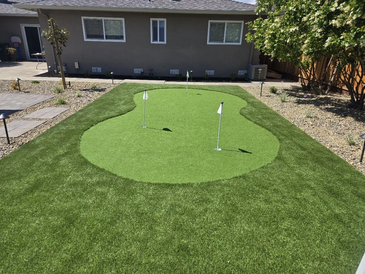Backyard putting green with three golf holes marked by flags, surrounded by neatly trimmed grass and gravel landscaping, with a house and trees in the background.