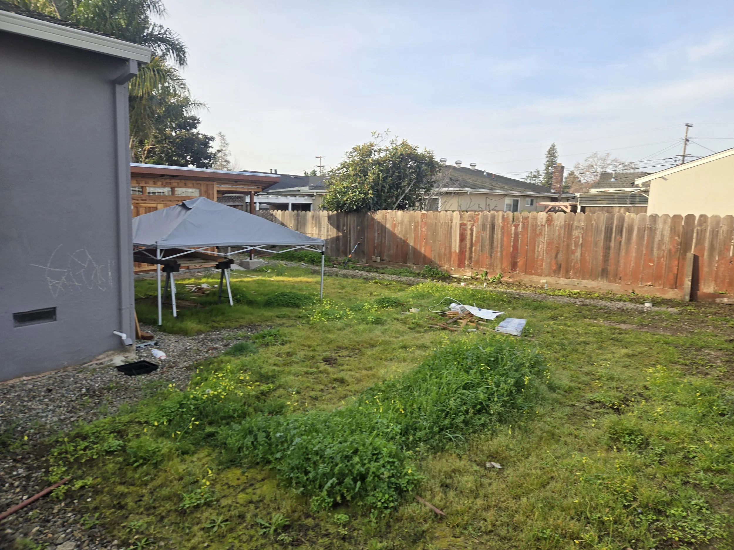 A backyard with a wooden fence, a small tree, a patio with a canopy, and neighboring houses under a cloudy sky.