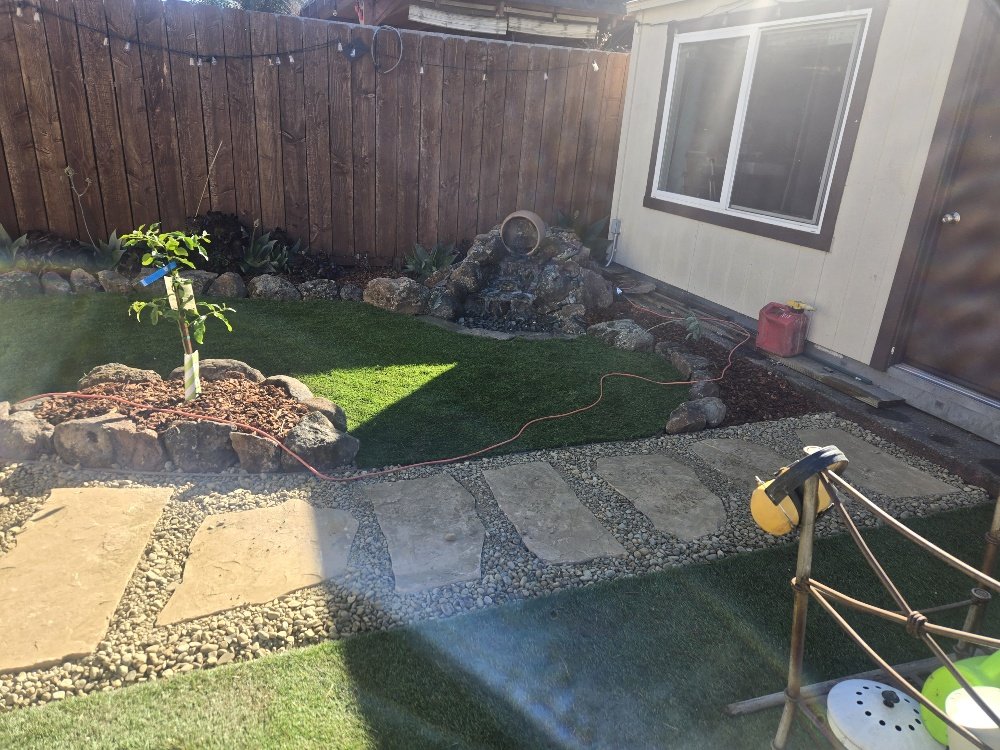 A backyard with a small artificial grass patch, a young tree planted in a mulched bed, a stone pathway, a water fountain feature with rocks, a wooden fence, a house with beige siding and a window, and some gardening or construction tools.